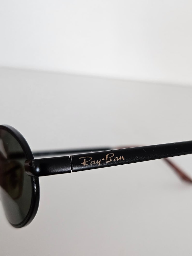 Puede incluir: Primer plano de gafas de sol Ray-Ban negras. Las gafas de sol tienen una montura redonda y lentes oscuras. El logotipo de Ray-Ban es visible en la patilla. La montura es negra con un acento marr&oacute;n en la patilla.