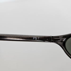 May include: Close-up of a pair of dark gray sunglasses. The text "PS 7" is printed in white on the arm of the sunglasses. The lenses are a dark green color.