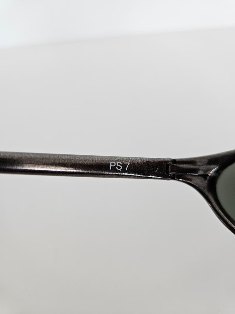 May include: Close-up of a pair of dark gray sunglasses. The text "PS 7" is printed in white on the arm of the sunglasses. The lenses are a dark green color.