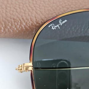 Puede incluir: Primer plano de gafas de sol Ray-Ban con montura dorada y lentes oscuras. El logotipo de Ray-Ban es visible en la lente superior derecha. Las gafas est&aacute;n parcialmente abiertas.