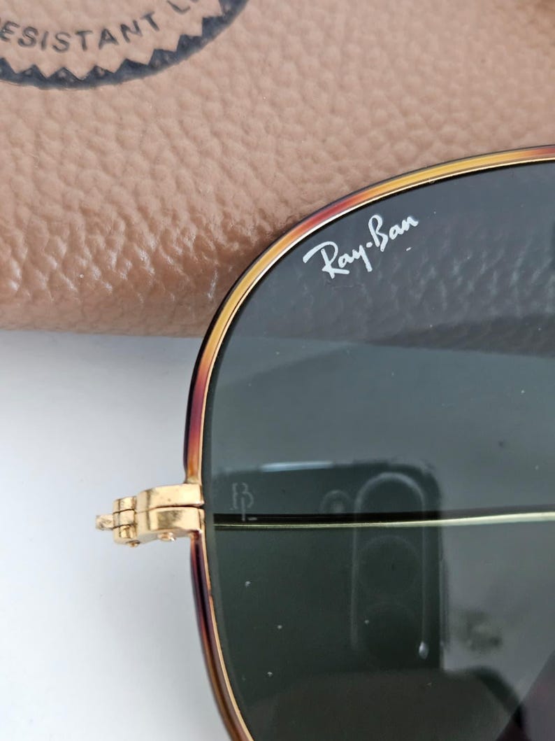 Puede incluir: Primer plano de gafas de sol Ray-Ban con montura dorada y lentes oscuras. El logotipo de Ray-Ban es visible en la lente superior derecha. Las gafas est&aacute;n parcialmente abiertas.