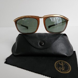 Pu&ograve; includere: Occhiali da sole Ray-Ban con barra frontale in metallo dorato, montature color marrone e lenti verde scuro. Il set include una custodia protettiva nera effetto pelle con chiusura a scatto e un panno per la pulizia nero con logo dorato.