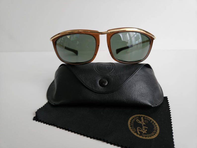 Pu&ograve; includere: Occhiali da sole Ray-Ban con barra frontale in metallo dorato, montature color marrone e lenti verde scuro. Il set include una custodia protettiva nera effetto pelle con chiusura a scatto e un panno per la pulizia nero con logo dorato.