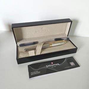 Op de afbeelding: Een zilver- en goudkleurige vulpen met een getextureerde houder ligt in een zwarte presentatiedoos met beige stoffen voering. De doos is aan de binnenzijde voorzien van het Sheaffer-logo. Twee inktpatronen en een Sheaffer-welkomstgids zijn inbegrepen.