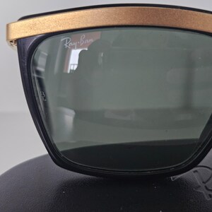 Puede incluir: Gafas de sol negras con lentes verdes y montura superior dorada. El logotipo de Ray-Ban es visible en la lente. Las gafas de sol est&aacute;n sobre una superficie negra.
