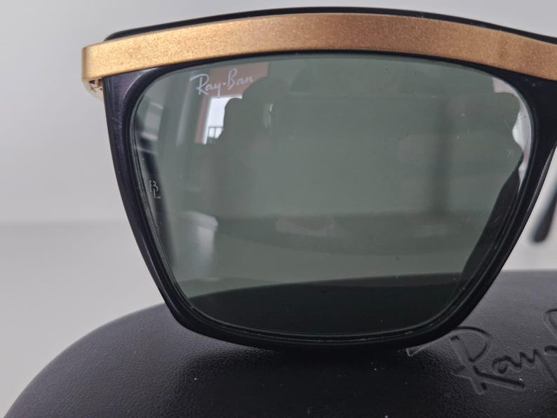 Puede incluir: Gafas de sol negras con lentes verdes y montura superior dorada. El logotipo de Ray-Ban es visible en la lente. Las gafas de sol est&aacute;n sobre una superficie negra.