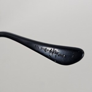 Puede incluir: Primer plano de una patilla de gafas negra con el texto "IL Ray-Ban USA" impreso en el interior. La patilla se curva ligeramente y tiene un acabado brillante, sobre un fondo blanco liso.