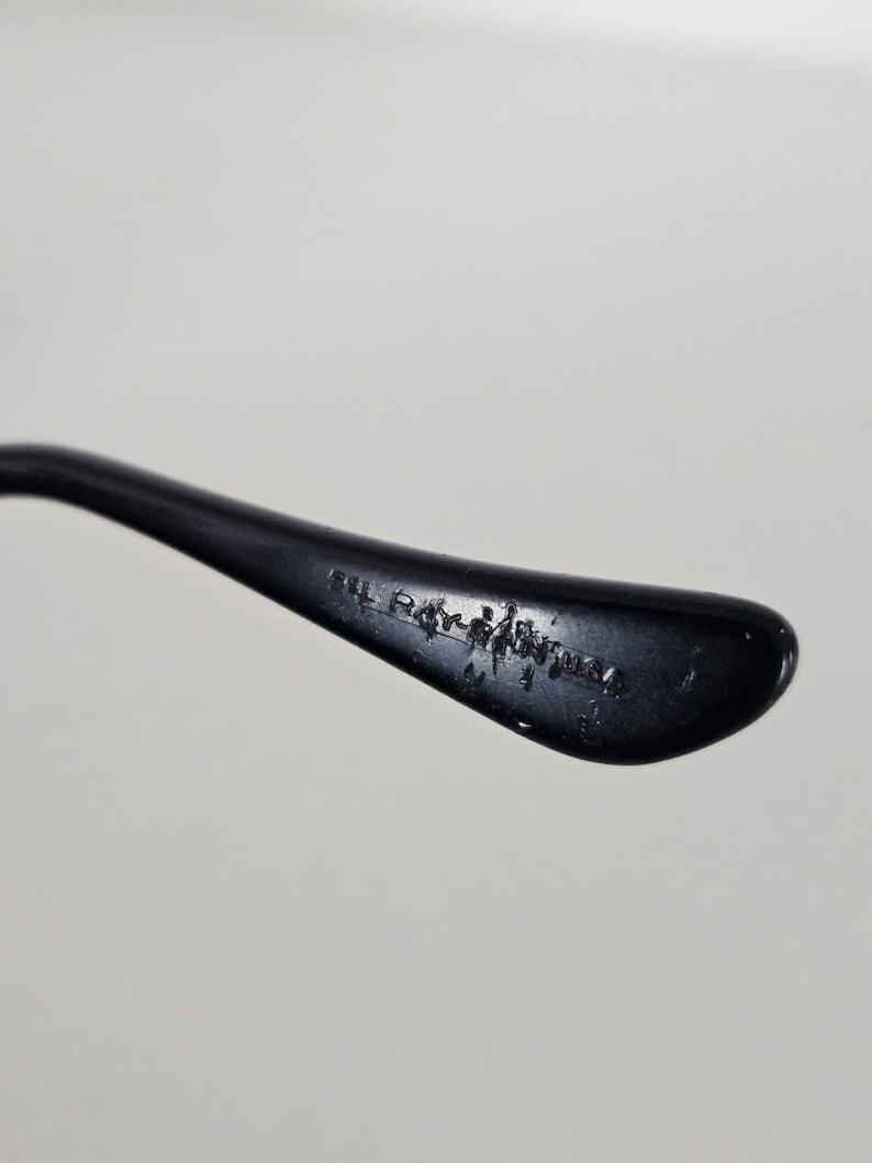 Puede incluir: Primer plano de una patilla de gafas negra con el texto "IL Ray-Ban USA" impreso en el interior. La patilla se curva ligeramente y tiene un acabado brillante, sobre un fondo blanco liso.