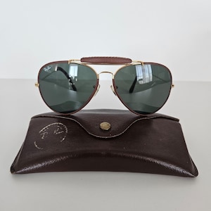 Puede incluir: Gafas de sol estilo aviador con puente y patillas de cuero marr&oacute;n, montura de metal dorado y lentes verde oscuro. Las gafas de sol descansan sobre un estuche de cuero marr&oacute;n con cierre de bot&oacute;n a presi&oacute;n. El estuche tiene un logotipo.