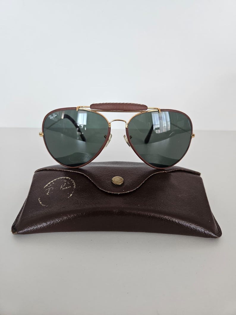 Puede incluir: Gafas de sol estilo aviador con puente y patillas de cuero marr&oacute;n, montura de metal dorado y lentes verde oscuro. Las gafas de sol descansan sobre un estuche de cuero marr&oacute;n con cierre de bot&oacute;n a presi&oacute;n. El estuche tiene un logotipo.