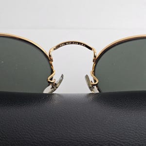 May include: Close-up of Ray-Ban sunglasses with gold frames and dark lenses. The bridge of the glasses is marked with "B&L RAY-BAN U.S.A." The sunglasses rest on a black textured surface.