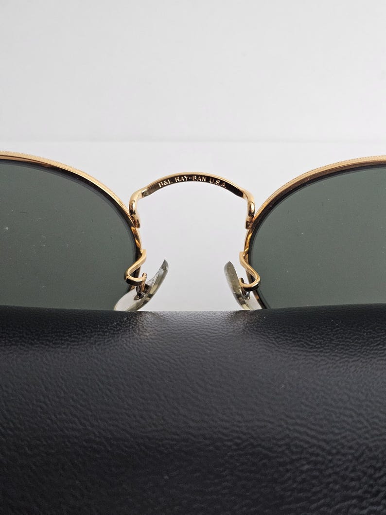 May include: Close-up of Ray-Ban sunglasses with gold frames and dark lenses. The bridge of the glasses is marked with "B&L RAY-BAN U.S.A." The sunglasses rest on a black textured surface.