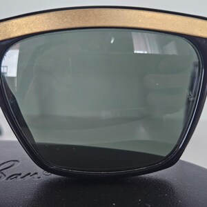 Puede incluir: Gafas de sol negras con montura superior dorada. Las lentes son de color verde oscuro. Las gafas de sol est&aacute;n sobre una superficie negra. El nombre de la marca es visible en la parte inferior de la montura.