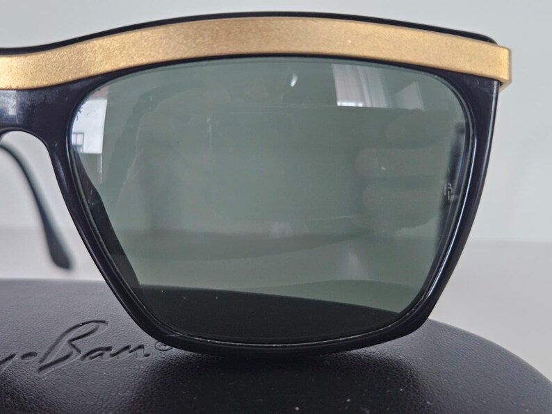 Puede incluir: Gafas de sol negras con montura superior dorada. Las lentes son de color verde oscuro. Las gafas de sol est&aacute;n sobre una superficie negra. El nombre de la marca es visible en la parte inferior de la montura.