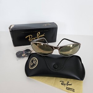 May include: Ray-Ban sunglasses with a silver frame and gold-tinted lenses, resting on a black leather case with a gold logo. The case is accompanied by a yellow cleaning cloth and a black box.