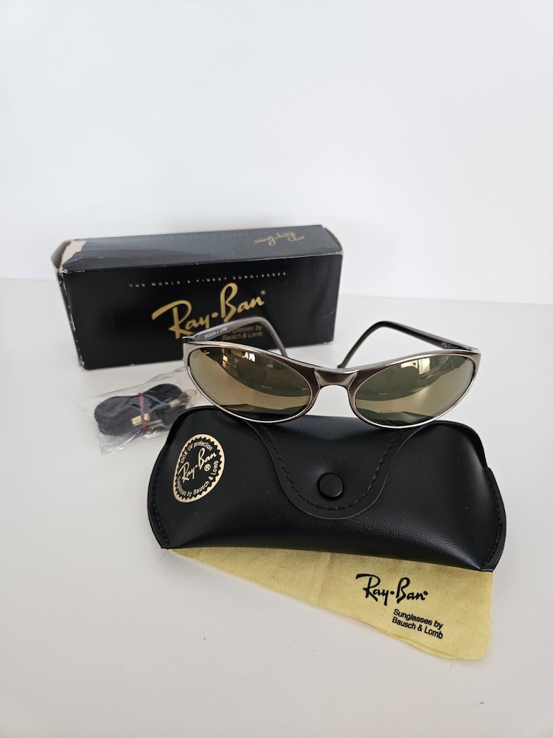 May include: Ray-Ban sunglasses with a silver frame and gold-tinted lenses, resting on a black leather case with a gold logo. The case is accompanied by a yellow cleaning cloth and a black box.