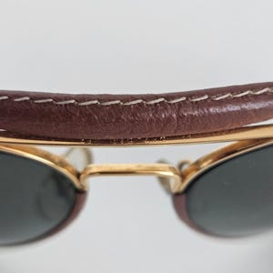Puede incluir: Primer plano de gafas de sol con una barra superior de cuero marr&oacute;n y monturas doradas. Las lentes son oscuras y la barra de cuero est&aacute; cosida con hilo blanco. Las gafas de sol est&aacute;n sobre un fondo blanco.