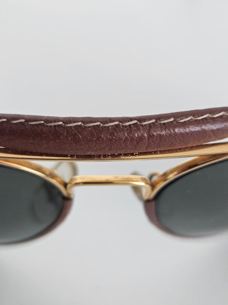 Puede incluir: Primer plano de gafas de sol con una barra superior de cuero marr&oacute;n y monturas doradas. Las lentes son oscuras y la barra de cuero est&aacute; cosida con hilo blanco. Las gafas de sol est&aacute;n sobre un fondo blanco.