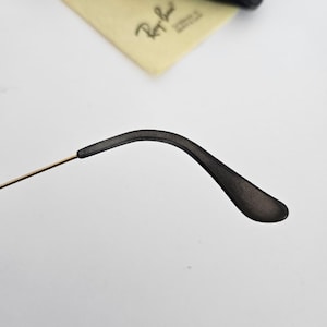 以下が含まれることがあります： 黒いメガネのテンプルアームのクローズアップ。金色のワイヤーと黒いイヤーピースが付いています。薄い黄色のクリーニングクロスと黒いケースが背景にあります。