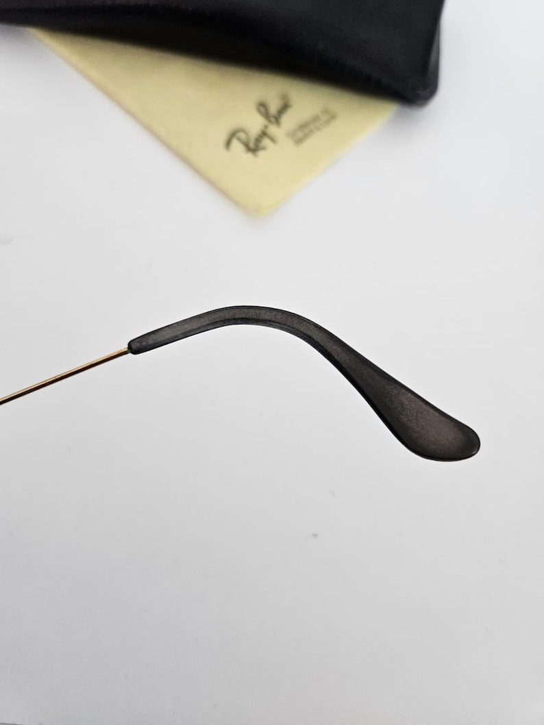 以下が含まれることがあります： 黒いメガネのテンプルアームのクローズアップ。金色のワイヤーと黒いイヤーピースが付いています。薄い黄色のクリーニングクロスと黒いケースが背景にあります。