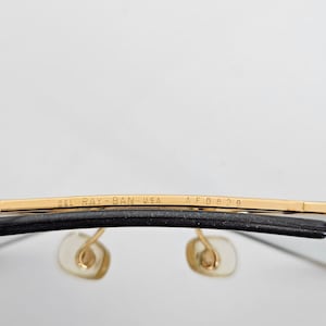 以下が含まれることがあります： レイバンのメガネのクローズアップ。上部のバーは金色で、「B&L RAY-BAN USA AF0829」の文字が入っています。レンズは透明で、ノーズパッドは薄いベージュ色です。フレームは黒色です。