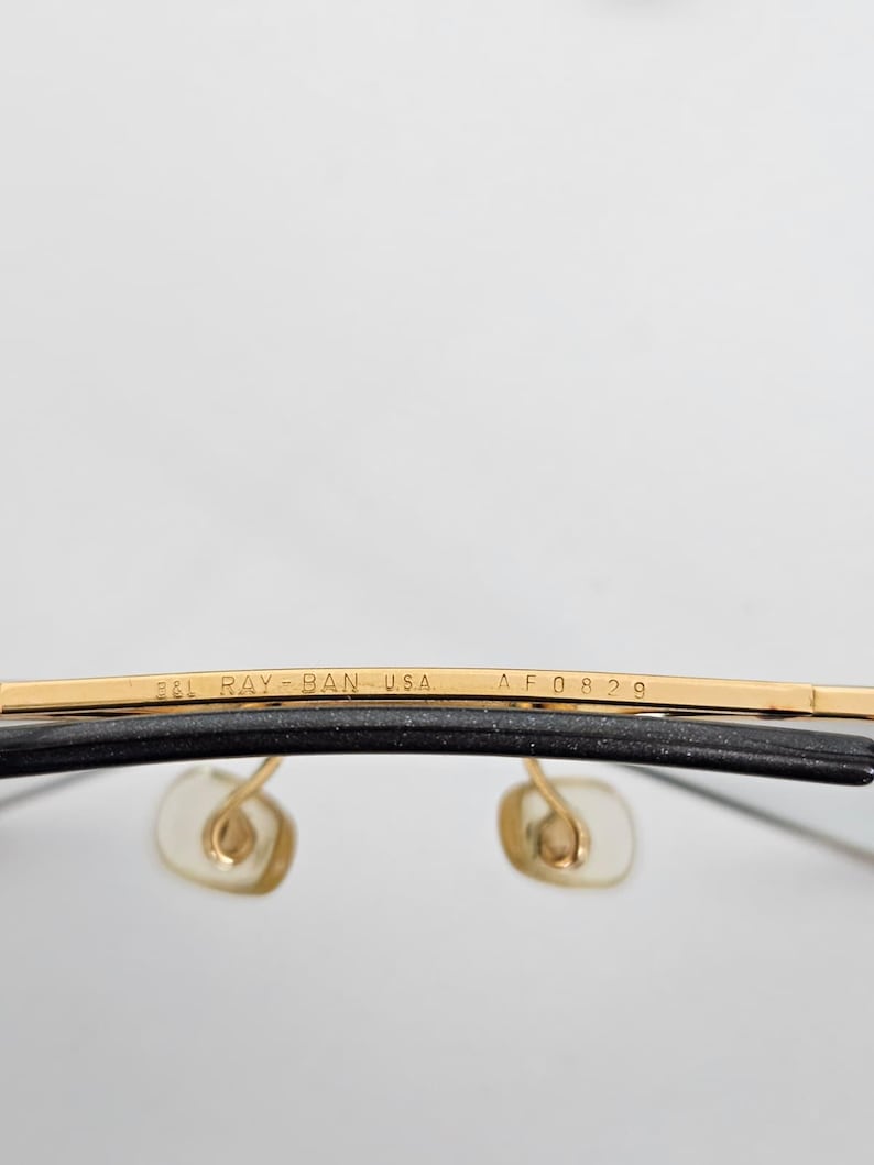 以下が含まれることがあります： レイバンのメガネのクローズアップ。上部のバーは金色で、「B&L RAY-BAN USA AF0829」の文字が入っています。レンズは透明で、ノーズパッドは薄いベージュ色です。フレームは黒色です。