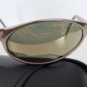 May include: Close-up of sunglasses with a metallic brown frame and green-tinted lenses. The sunglasses are resting on a black leather case. The lenses reflect the surrounding environment.