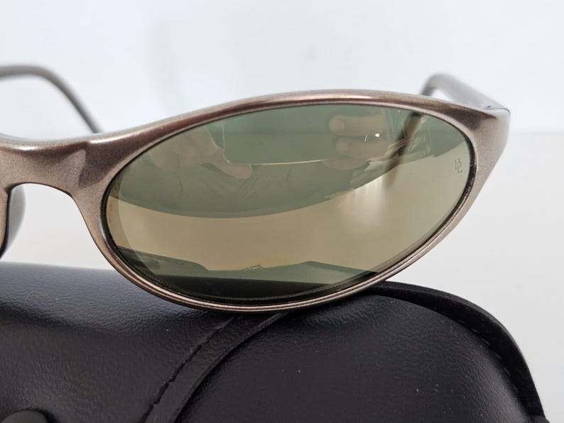 May include: Close-up of sunglasses with a metallic brown frame and green-tinted lenses. The sunglasses are resting on a black leather case. The lenses reflect the surrounding environment.