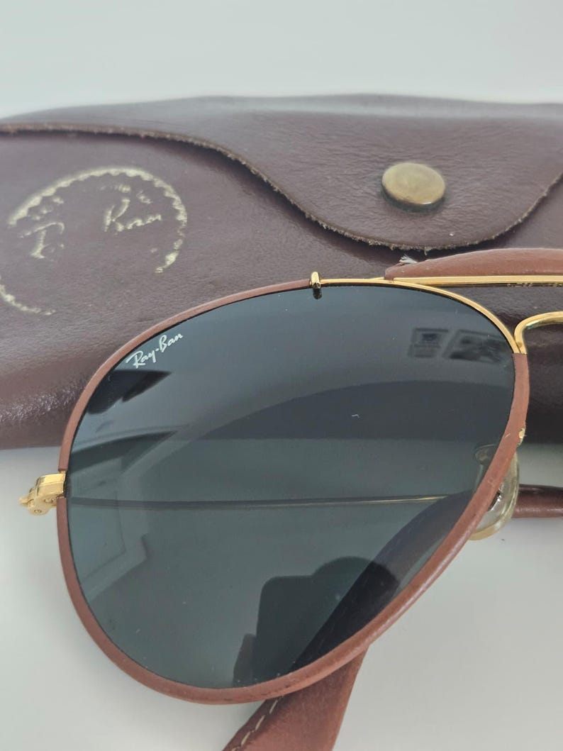 Puede incluir: Gafas de sol de cuero marr&oacute;n con lentes oscuras y montura dorada. Las gafas de sol est&aacute;n en un estuche de cuero marr&oacute;n con el logotipo de Ray-Ban. El estuche tiene un cierre de bot&oacute;n.