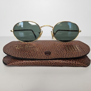 May include: Gold-rimmed sunglasses with oval lenses sit on a brown leather case. The sunglasses have dark green lenses and gold arms. The case is textured and has a button closure. The sunglasses and case are on a white surface.