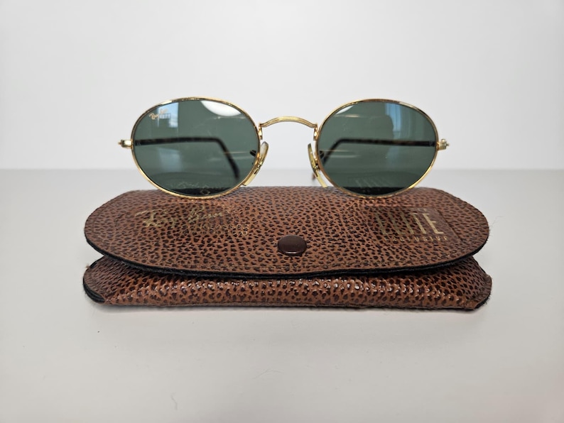 May include: Gold-rimmed sunglasses with oval lenses sit on a brown leather case. The sunglasses have dark green lenses and gold arms. The case is textured and has a button closure. The sunglasses and case are on a white surface.