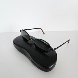 Puede incluir: Gafas de sol ovaladas negras con lentes oscuras y monturas negras finas, colocadas sobre un estuche negro para gafas de sol. Las patillas tienen puntas marrones. El conjunto est&aacute; sobre un fondo blanco.
