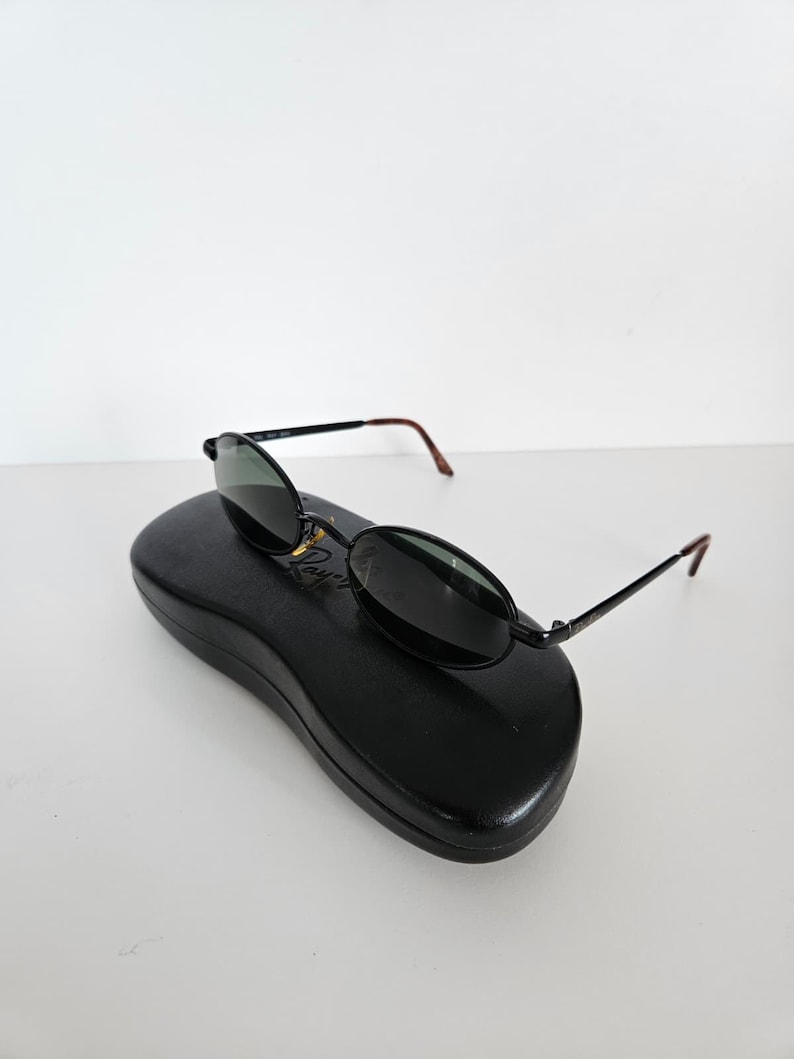 Puede incluir: Gafas de sol ovaladas negras con lentes oscuras y monturas negras finas, colocadas sobre un estuche negro para gafas de sol. Las patillas tienen puntas marrones. El conjunto est&aacute; sobre un fondo blanco.