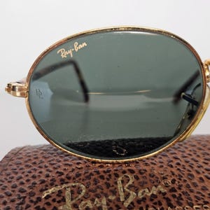 May include: Gold-rimmed Ray-Ban sunglasses with green lenses. The Ray-Ban logo is visible on the top left lens. The sunglasses are resting on a brown leather case with the Ray-Ban logo embossed on it.