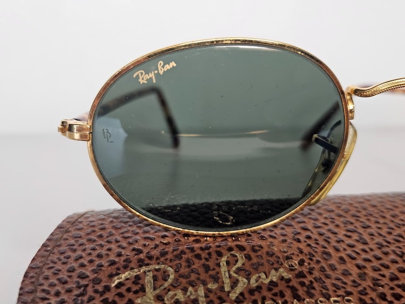 May include: Gold-rimmed Ray-Ban sunglasses with green lenses. The Ray-Ban logo is visible on the top left lens. The sunglasses are resting on a brown leather case with the Ray-Ban logo embossed on it.