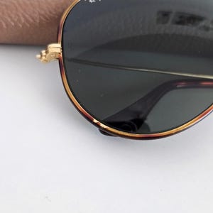 Puede incluir: Primer plano de gafas de sol Ray-Ban con montura dorada y lentes verde oscuro. Las gafas de sol tienen una forma de aviador cl&aacute;sica con un patr&oacute;n de carey en la montura. El logotipo de Ray-Ban es visible en la lente.