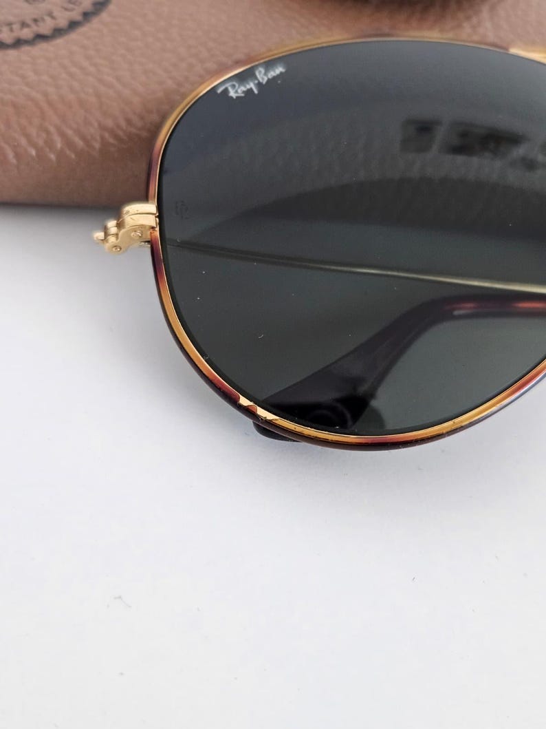Puede incluir: Primer plano de gafas de sol Ray-Ban con montura dorada y lentes verde oscuro. Las gafas de sol tienen una forma de aviador cl&aacute;sica con un patr&oacute;n de carey en la montura. El logotipo de Ray-Ban es visible en la lente.