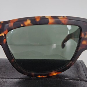 May include: Close-up of sunglasses with a tortoiseshell pattern frame and green lenses. The sunglasses are resting on a black surface. The frame has a dark brown base with orange and yellow accents.