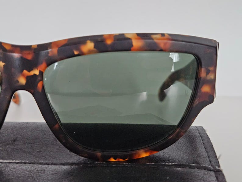 May include: Close-up of sunglasses with a tortoiseshell pattern frame and green lenses. The sunglasses are resting on a black surface. The frame has a dark brown base with orange and yellow accents.