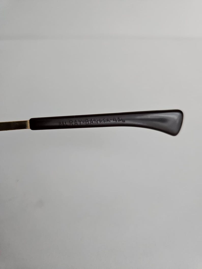 Pu&ograve; includere: Un'asta di occhiali marrone scuro con una cerniera in metallo color oro. Il lato interno del terminale presenta il testo in rilievo B&L RAY-BAN U.S.A. 43/14, che indica il marchio e le misure specifiche della montatura degli occhiali.