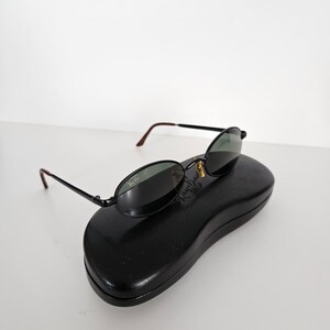 Puede incluir: Gafas de sol ovaladas negras con lentes verde oscuro y montura negra, sobre un estuche negro para gafas de sol. Las gafas tienen un detalle dorado y el logotipo de Ray-Ban es visible en la lente.