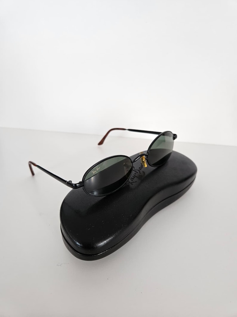 Puede incluir: Gafas de sol ovaladas negras con lentes verde oscuro y montura negra, sobre un estuche negro para gafas de sol. Las gafas tienen un detalle dorado y el logotipo de Ray-Ban es visible en la lente.