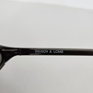 May include: Close-up of a dark-colored eyeglass frame with the text "BAUSCH & LOMB" printed in white. The frame is set against a plain white background.