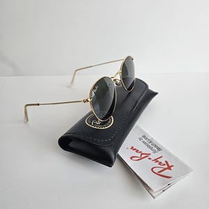 May include: Gold-framed sunglasses with dark lenses rest on a black leather case. The case has a gold emblem. A folded white paper with red text is next to the case. The sunglasses have a classic, round shape.