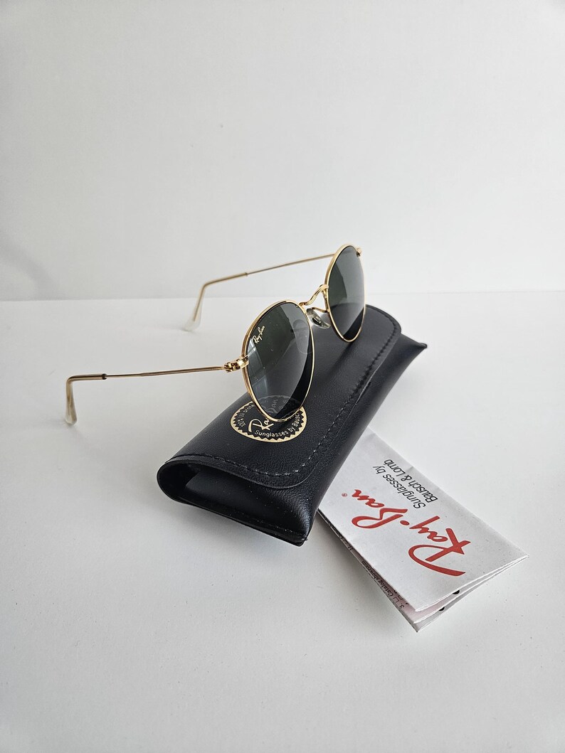 May include: Gold-framed sunglasses with dark lenses rest on a black leather case. The case has a gold emblem. A folded white paper with red text is next to the case. The sunglasses have a classic, round shape.