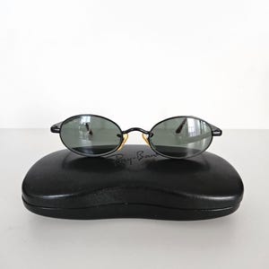 Puede incluir: Gafas de sol ovaladas negras con lentes oscuras sobre un estuche negro. Las gafas tienen una montura negra fina y detalles dorados. El estuche es rectangular y tiene el logotipo de Ray-Ban.