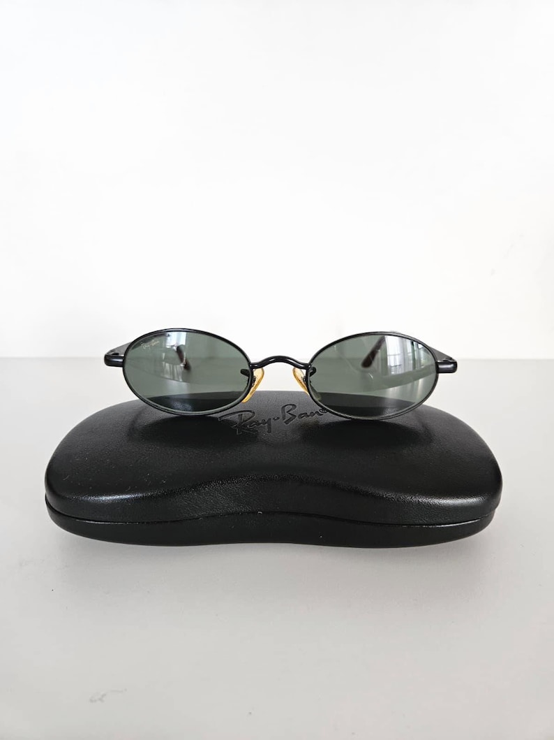Puede incluir: Gafas de sol ovaladas negras con lentes oscuras sobre un estuche negro. Las gafas tienen una montura negra fina y detalles dorados. El estuche es rectangular y tiene el logotipo de Ray-Ban.