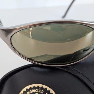May include: Close-up of a pair of silver-framed sunglasses with reflective lenses. The sunglasses are resting on a black case with a gold emblem. The lenses have a slight green tint.