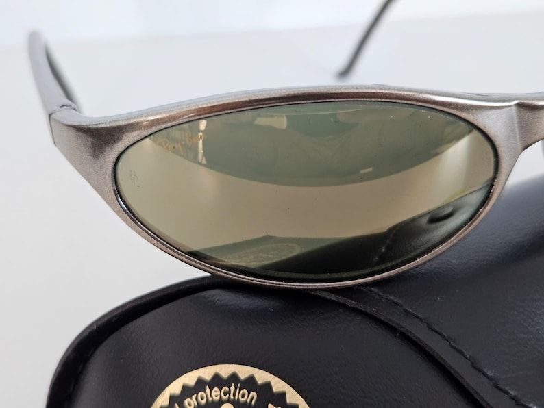 May include: Close-up of a pair of silver-framed sunglasses with reflective lenses. The sunglasses are resting on a black case with a gold emblem. The lenses have a slight green tint.