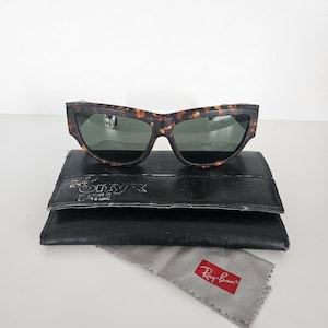 May include: Tortoiseshell-patterned sunglasses with green lenses rest on a black leather case. A gray cleaning cloth with a red Ray-Ban logo is below. The sunglasses have a distinctive, angular frame design.